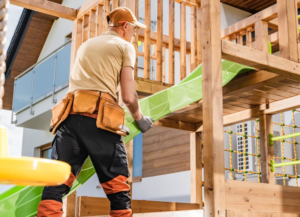 A man is engaged in installing a vibrant green slide onto a wooden playground structure in a residential backyard. Wearing a cap and work gloves, he is focused on ensuring the slide is securely attached.