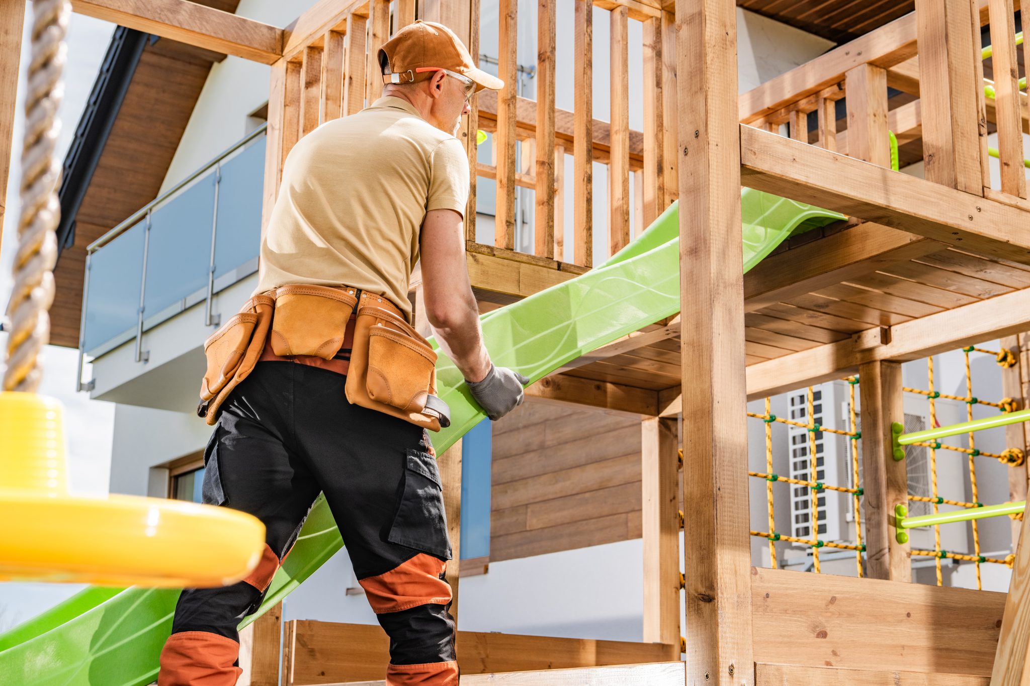 A man is engaged in installing a vibrant green slide onto a wooden playground structure in a residential backyard. Wearing a cap and work gloves, he is focused on ensuring the slide is securely attached.